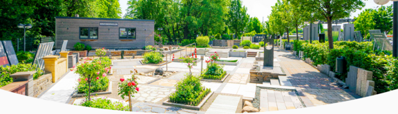 Außenaufnahme des bauwelt-Standorts in Norderstedt mit einem weitläufigen Ausstellungspark für Garten- und Landschaftsbau. Pflasterflächen, Mauersysteme, Pflanzbeete und moderne Gestaltungselemente sind harmonisch angeordnet. Im Hintergrund steht ein Holzpavillon mit der Aufschrift „Beratungspavillon“, umgeben von Grünflächen, Bäumen und blühenden Rosenbeeten.
