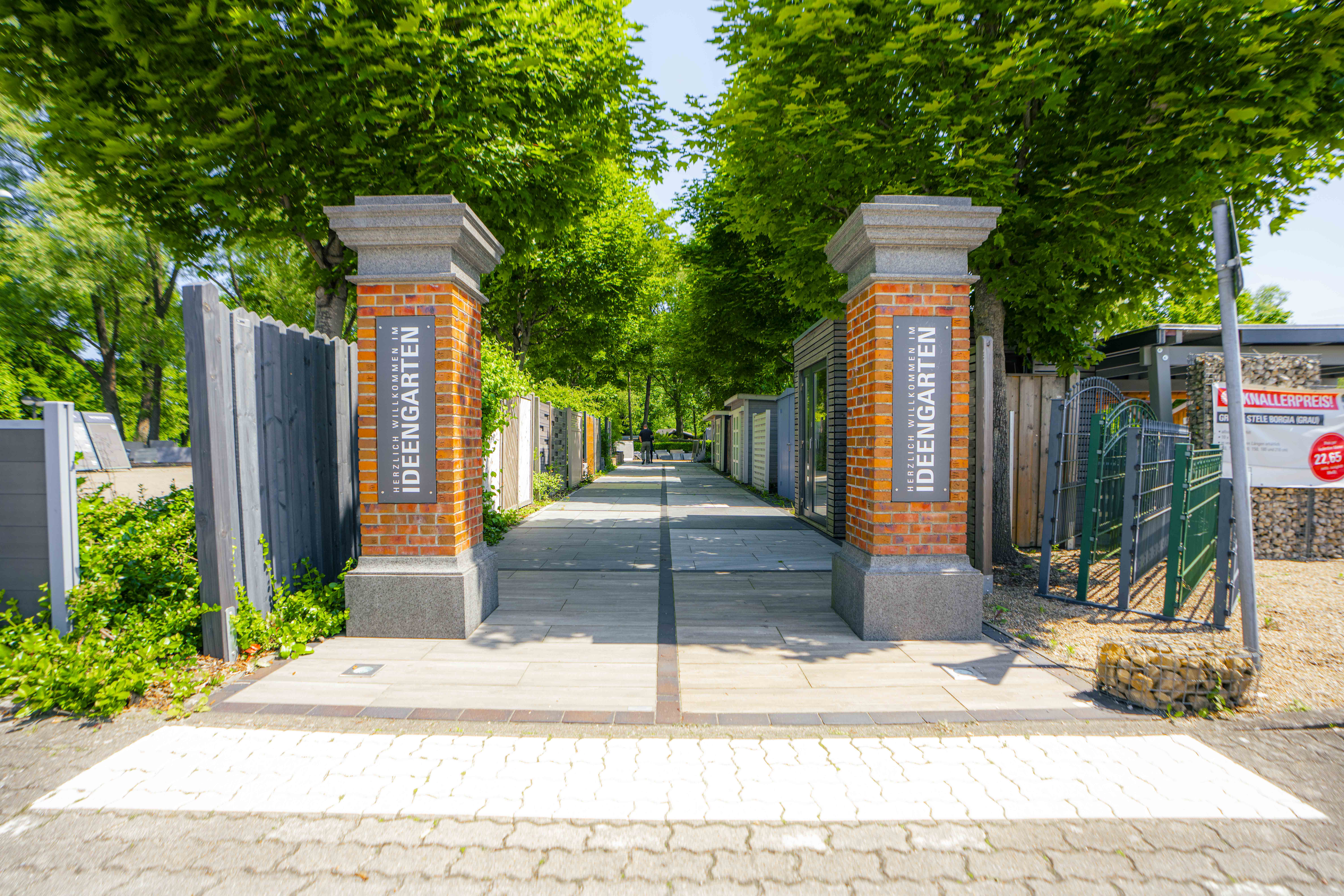 Eingang mit zwei gemauerten Pfeilern und Schildern „Herzlich willkommen im Ideengarten“, flankiert von Bäumen und Ausstellungsflächen für Gartenprodukte.