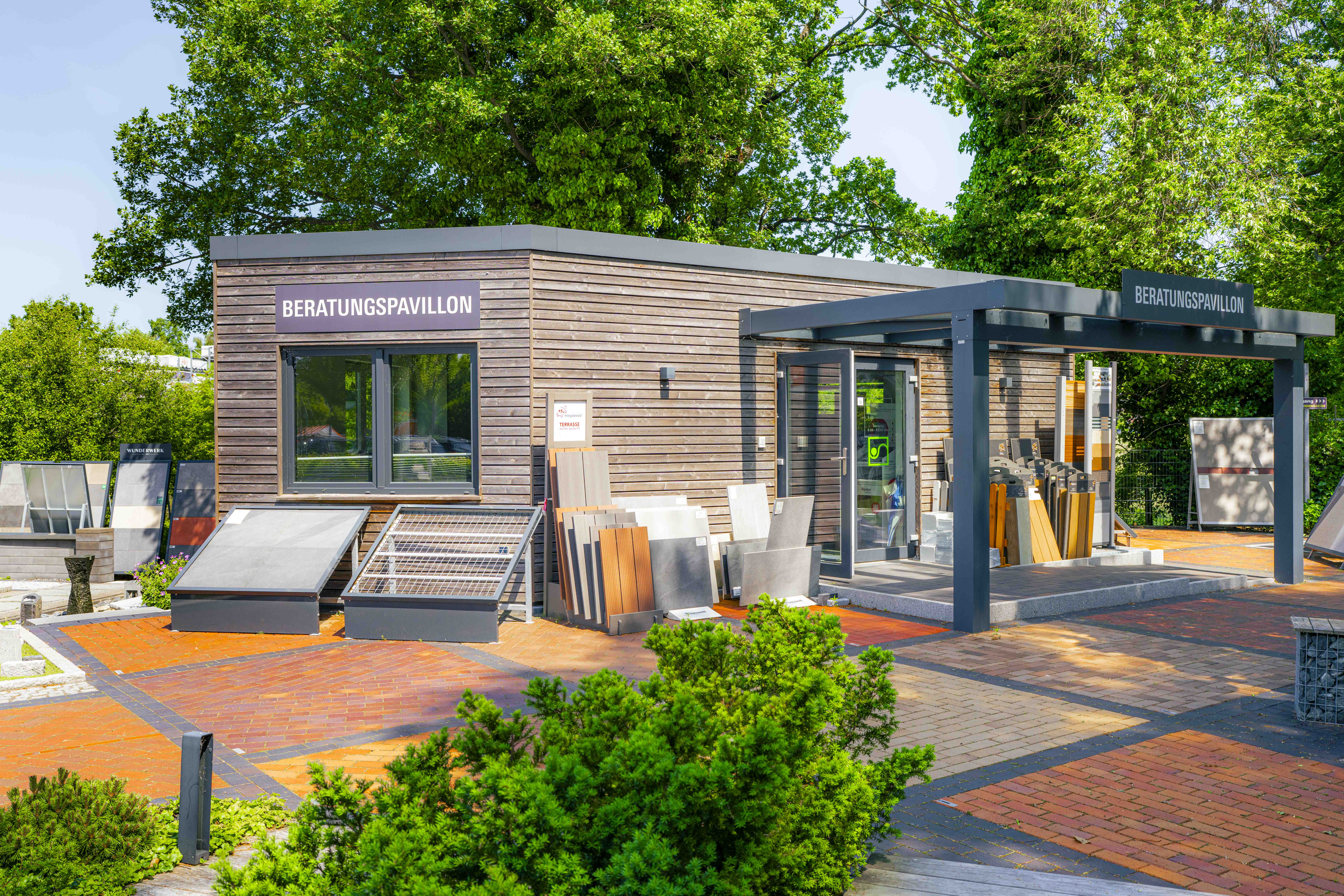 Beratungs-Pavillon aus Holz mit großen Fenstern, davor Musterflächen mit Platten und Terrassenbelägen.
