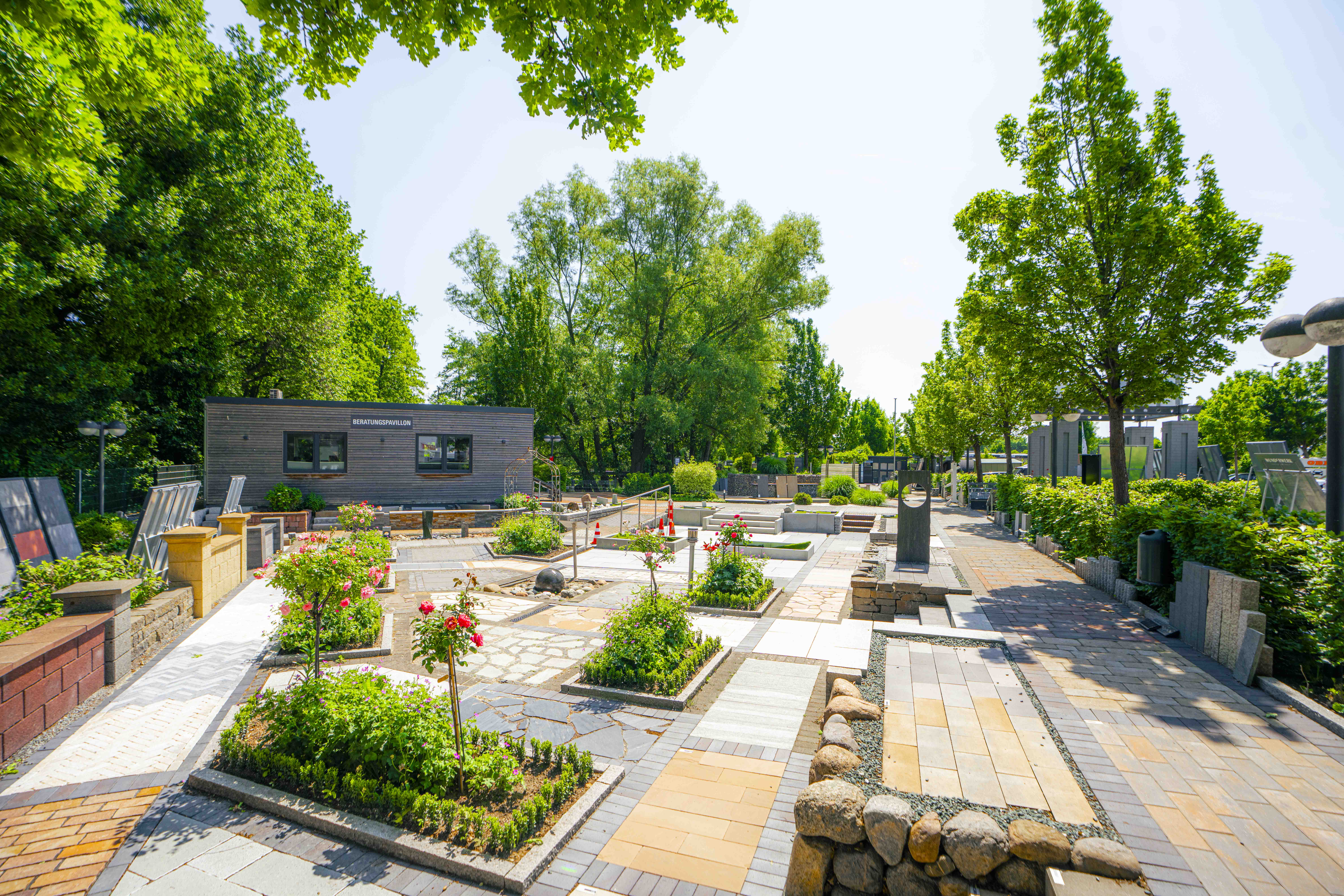 Musterfläche im Ideengarten mit Pflastersteinen, Wegen, kleinen Beeten und Skulpturen, im Hintergrund der Beratungs-Pavillon.