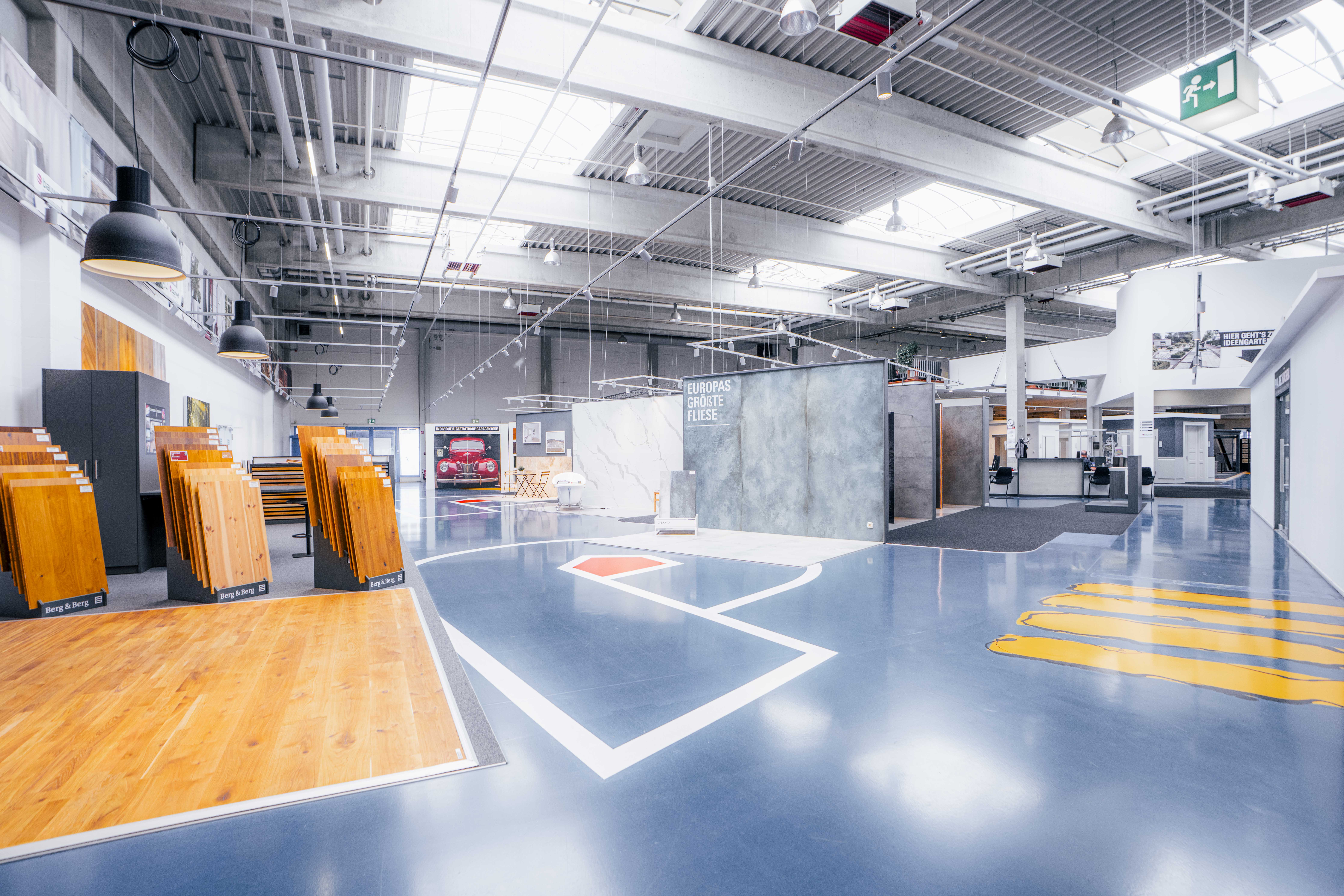 Innenansicht eines Baumarkts mit Farbeimern, Werkzeugen, Bauzubehör und Regalen, gelber und blauer Boden.